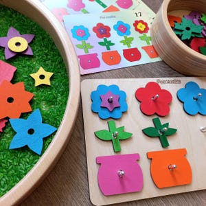 May include: A wooden educational toy with a flower theme. The toy has a base with six holes and six colorful flower pieces with pegs that fit into the holes. The flower pieces are blue, red, green, and pink. The toy is labeled "Poznavalka" which is Polish for "Cognitive".