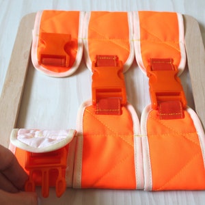 Puede incluir: Correas de tela naranja con hebillas de pl&aacute;stico naranja unidas a un marco de madera. Las correas est&aacute;n forradas con tela blanca y tienen un patr&oacute;n acolchado.
