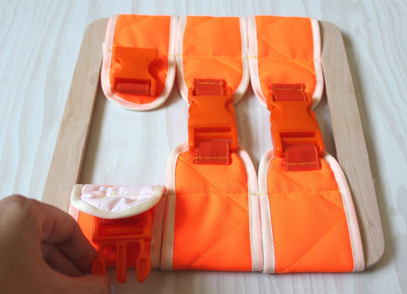 Puede incluir: Correas de tela naranja con hebillas de pl&aacute;stico naranja unidas a un marco de madera. Las correas est&aacute;n forradas con tela blanca y tienen un patr&oacute;n acolchado.