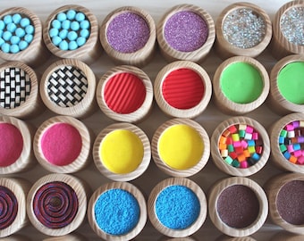 May include: A set of 16 wooden sensory toys with different textures and colours. Each toy has a circular wooden base with a different material inside, such as glitter, beads, sand, and fabric.
