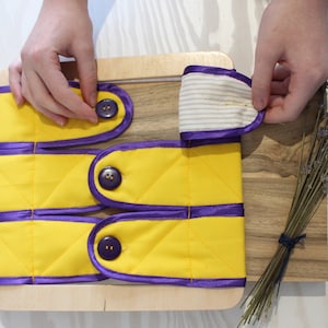 Puede incluir: Una tabla de madera con tres tiras de tela amarilla con ribete morado y botones. Cada tira tiene dos botones. Las tiras de tela est&aacute;n dobladas y los botones son visibles. Una mano se muestra sosteniendo una de las tiras de tela.