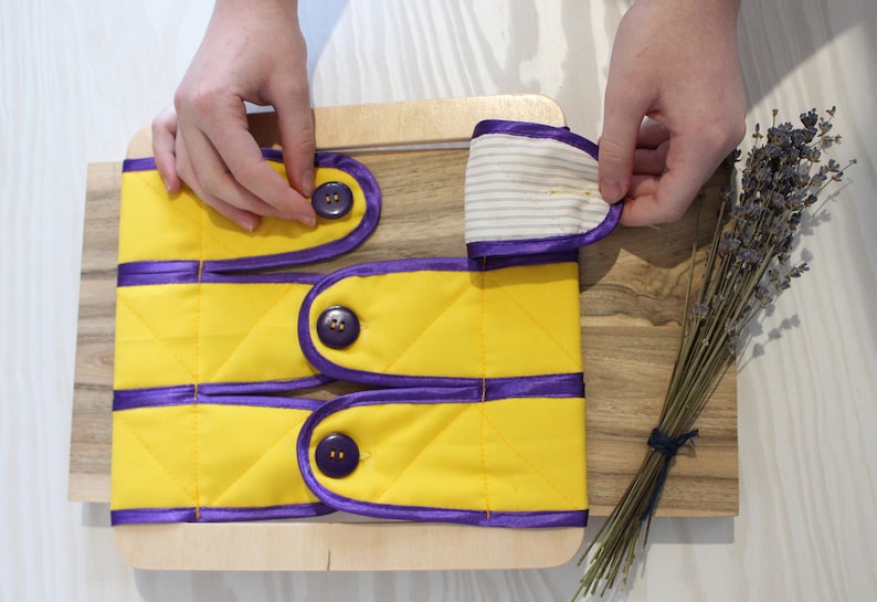 Puede incluir: Una tabla de madera con tres tiras de tela amarilla con ribete morado y botones. Cada tira tiene dos botones. Las tiras de tela est&aacute;n dobladas y los botones son visibles. Una mano se muestra sosteniendo una de las tiras de tela.