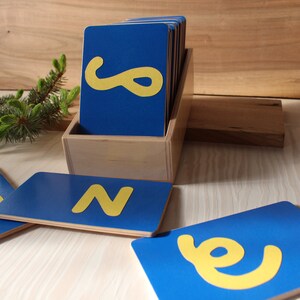May include: A set of blue wooden alphabet letter tiles with yellow letters. The tiles are in a wooden box. The letters are N and e.