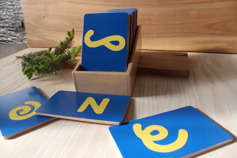 May include: A set of blue wooden alphabet letter tiles with yellow letters. The tiles are in a wooden box. The letters are N and e.