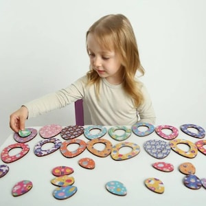 May include: A child plays with a set of colorful wooden Easter egg puzzle pieces. The pieces are decorated with various patterns and designs. The child is holding one piece in their hand and is looking at the other pieces on the table.