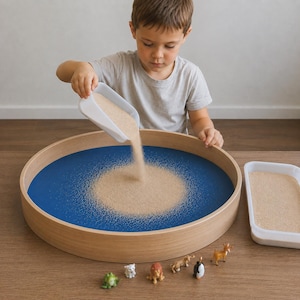May include: A child pours sand from a white scoop into a large, round wooden tray with a blue interior. Small animal figurines are arranged at the bottom of the tray. A second tray filled with sand is on the right.