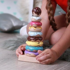 Puede incluir: Un juguete de apilar de madera de donut colorido con nueve piezas, que incluyen un donut con cobertura de chocolate, un donut glaseado rosa, un donut glaseado amarillo, un donut glaseado azul, un donut glaseado marrón, un donut glaseado amarillo, un donut glaseado turquesa, un donut glaseado morado y una base de madera.