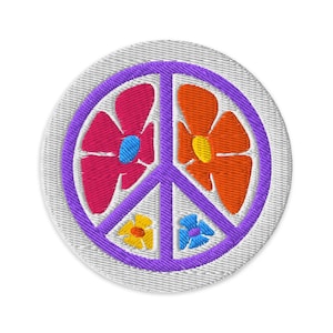Könnte beinhalten: Gestickter Aufnäher mit einem lila Friedenssymbol und bunten Blumen. Die Blumen sind rosa, orange, gelb und blau, vor einem weißen, strukturierten Hintergrund. Ein lebendiger, dekorativer Aufnäher.