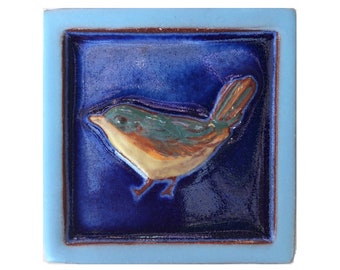 May include: A blue ceramic tile with a raised image of a brown, green, and yellow bird on a dark blue background.
