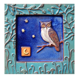 May include: A blue ceramic tile with a raised design of an owl perched on a branch with a crescent moon in the lower left corner. The tile is surrounded by a decorative border of leaves and vines.