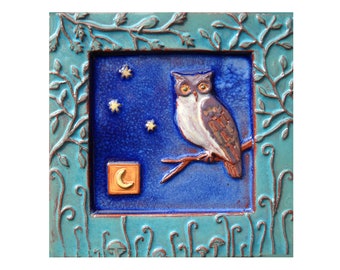 May include: A blue ceramic tile with a raised design of an owl perched on a branch with a crescent moon in the lower left corner. The tile is surrounded by a decorative border of leaves and vines.