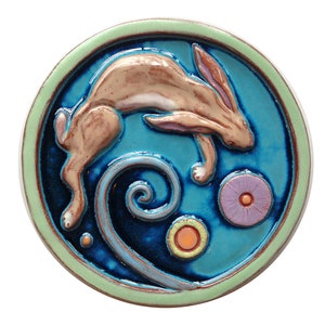May include: A ceramic tile with a blue background and a brown rabbit jumping over a blue swirl. The tile is surrounded by a green border and has two small orange and purple flowers.
