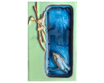 May include: A ceramic tile with a light green background and a blue and white bird perched on a branch inside a blue rectangular frame. The frame is surrounded by a brown border. There are small yellow flowers on a brown branch to the left of the frame.