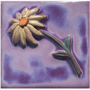 May include: A ceramic tile with a raised design of a yellow daisy with an orange center and green leaves on a purple background.