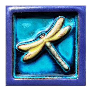 May include: A ceramic tile with a raised, yellow dragonfly design on a blue background. The tile is framed with a purple border.
