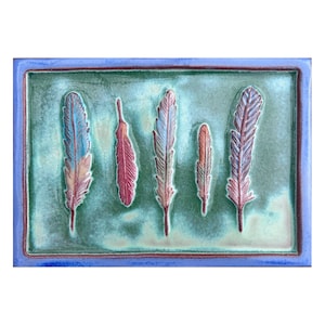 May include: A rectangular ceramic tile with a blue border and a green background. Five stylized feathers are embossed on the tile in shades of blue, red, and pink.