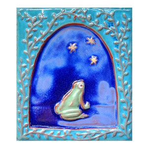 May include: A ceramic tile with a blue and purple background and a green frog looking up at three gold stars. The tile is framed with a turquoise border with silver vines.