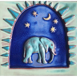 May include: A blue and green ceramic tile with a raised design of an elephant standing under a crescent moon and stars. The tile has a textured border of blue and green triangles.