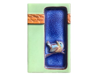 May include: A light green ceramic tile with a blue and white bird on a branch in a blue rectangle. The tile has a brown and orange border.
