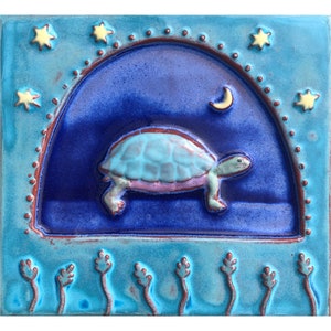 May include: A blue ceramic tile with a 3D image of a turtle walking under a crescent moon and stars. The tile has a blue and purple background with a brown border.