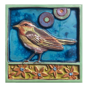 May include: A ceramic tile with a blue background and a brown bird with yellow accents. The bird is perched on a ledge with orange flowers. There are two blue and purple circles in the background.