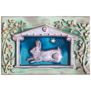 May include: A ceramic tile with a green background and a pink rabbit in a blue frame. The rabbit is under a pink roof with a crescent moon and a star. The tile is surrounded by green leaves and yellow flowers.