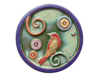 May include: A round ceramic tile with a green background and a colorful bird with red, yellow, and orange accents. The tile is decorated with swirls and circles in shades of blue, orange, and yellow.