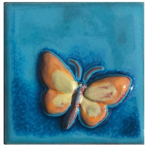 May include: Square ceramic tile with a raised butterfly design. The butterfly has orange and yellow wings with a blue body, set against a turquoise background. The tile has a glossy finish and a textured appearance.