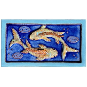 May include: A blue ceramic tile with a raised design of two gold fish swimming in a circle. The tile has a light blue border.