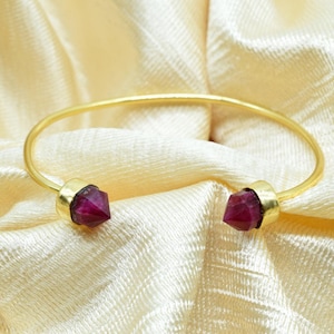 Puede incluir: Un brazalete dorado con un diseño sencillo. El brazalete presenta dos gemas facetadas de color rojo intenso en cada extremo. El brazalete se exhibe sobre una tela crema texturizada.