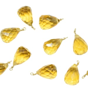 May include: A collection of faceted, teardrop-shaped citrine gemstone pendants with gold-colored wire loops. The pendants are a warm yellow color and are scattered against a white background. These pendants are suitable for jewelry making.