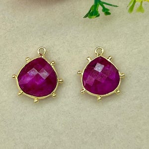 May include: Two magenta-colored, teardrop-shaped pendants with gold-colored metal trim. The pendants have a faceted surface and a small loop at the top for attaching to a chain or earring hook. The background includes pink and white flowers.