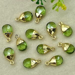 May include: A selection of green, teardrop-shaped beads with gold-coloured caps and loops. The faceted beads are arranged on a light beige surface, with some greenery and flowers in the background.