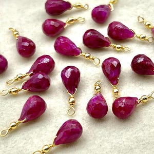 Könnte beinhalten: Eine Sammlung tropfenförmiger, facettierter, tiefmagentafarbener Edelsteinanhänger mit goldfarbenen Drahtschlaufen. Die Anhänger sind auf einer hellen Oberfläche verstreut, mit ein paar rosa Blumen im Hintergrund.