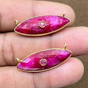 Könnte beinhalten: Zwei ovale, magentafarbene Edelsteinanhänger mit goldfarbenem Metallbesatz und kleinen Schlaufen an jedem Ende. Jeder Anhänger hat einen kleineren, kreisförmigen rosa Stein in der Mitte. Die Anhänger werden vor einem neutralen Hintergrund präsentiert.