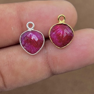 May include: Two heart-shaped ruby pendants. One pendant has a silver bezel, and the other has a gold bezel. Both pendants have a small loop at the top for a chain. The rubies are a deep red color.