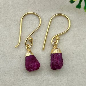 Op de afbeelding: Een paar goudkleurige oorhangers met ruwe, diep magenta robijn edelstenen. De robijnstenen zijn afgedekt met goud en hangen aan goudkleurige oorhaken. De oorbellen worden gepresenteerd op een lichtgrijze ondergrond.