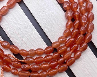 Puede incluir: Cadenas de cuentas ovaladas de color marrón anaranjado, probablemente piedras preciosas, dispuestas en espiral. Las cuentas están ensartadas en un fino cordón naranja a juego. El fondo es una superficie de madera clara con rayas negras.