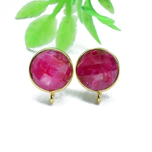 Pode incluir: Um par de brincos dourados com pedras redondas de rubi vermelho facetadas. Os brincos têm um design simples e elegante.