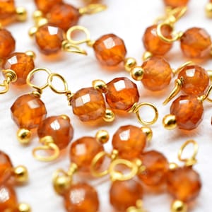 Puede incluir: Primer plano de un grupo de cuentas de piedras preciosas de color naranja con anillos de salto dorados. Las cuentas son facetadas y tienen un acabado brillante.