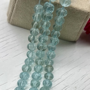 Könnte beinhalten: Stränge hellblauer Edelsteinperlen mit einem geschnitzten, gerippten Design. Die Perlen sind aufgefädelt und bilden eine Halskette oder ein Armband. Die Perlen werden auf einer beigen Box mit weißem Hintergrund präsentiert.
