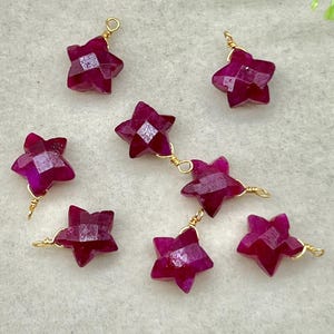 Puede incluir: Primer plano de varias gemas facetadas en forma de estrella, de un tono magenta intenso. Cada estrella está cableada individualmente con alambre dorado, algunas conectadas en cadena. Las gemas están dispuestas sobre una superficie clara.