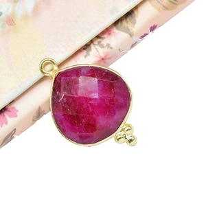 May include: A teardrop-shaped pendant with a deep red gemstone set in gold. The pendant has two small gold beads on the side.