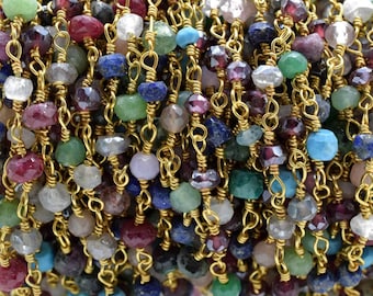 May include: A close-up of a gold chain with various coloured gemstone beads. The beads are small and round, and they are strung together in a random pattern. The chain is made of gold-coloured metal and is about 6 mm wide.