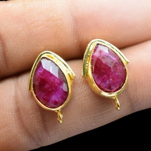 May include: A pair of gold-tone earrings with red gemstone drops. The earrings have a teardrop shape and are set with a single red gemstone in each.