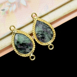 May include: Two gold-toned teardrop-shaped pendants with green and black gemstone inlays. Each pendant has a loop on either side for attaching to a chain or cord.