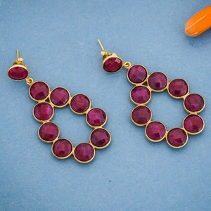 Puede incluir: Un par de pendientes colgantes con forma de lágrima. Los pendientes están hechos de metal dorado y presentan un círculo de piedras preciosas redondas, facetadas y de color rosa oscuro.