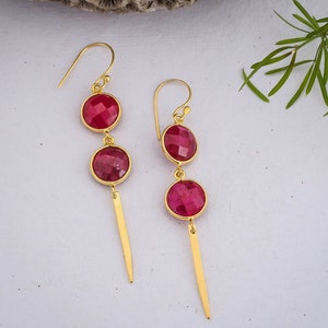 May include: A pair of gold earrings with three red gemstone drops. The earrings have a long, thin, gold dangle.