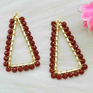 Pode incluir: Um par de brincos triangulares de ouro com contas vermelhas. Os brincos estão sobre um fundo branco.