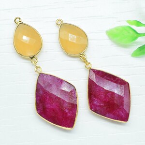 Könnte beinhalten: Ein Paar Hängeohrringe mit einem tropfenförmigen gelben Edelstein und einem größeren, facettierten, rosafarbenen Edelstein. Die Ohrringe haben goldfarbene Metallfassungen.